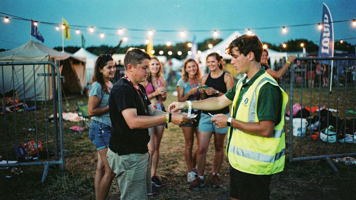 Group leader checking wristbands for young travellers at a festival campsite entrance at dusk